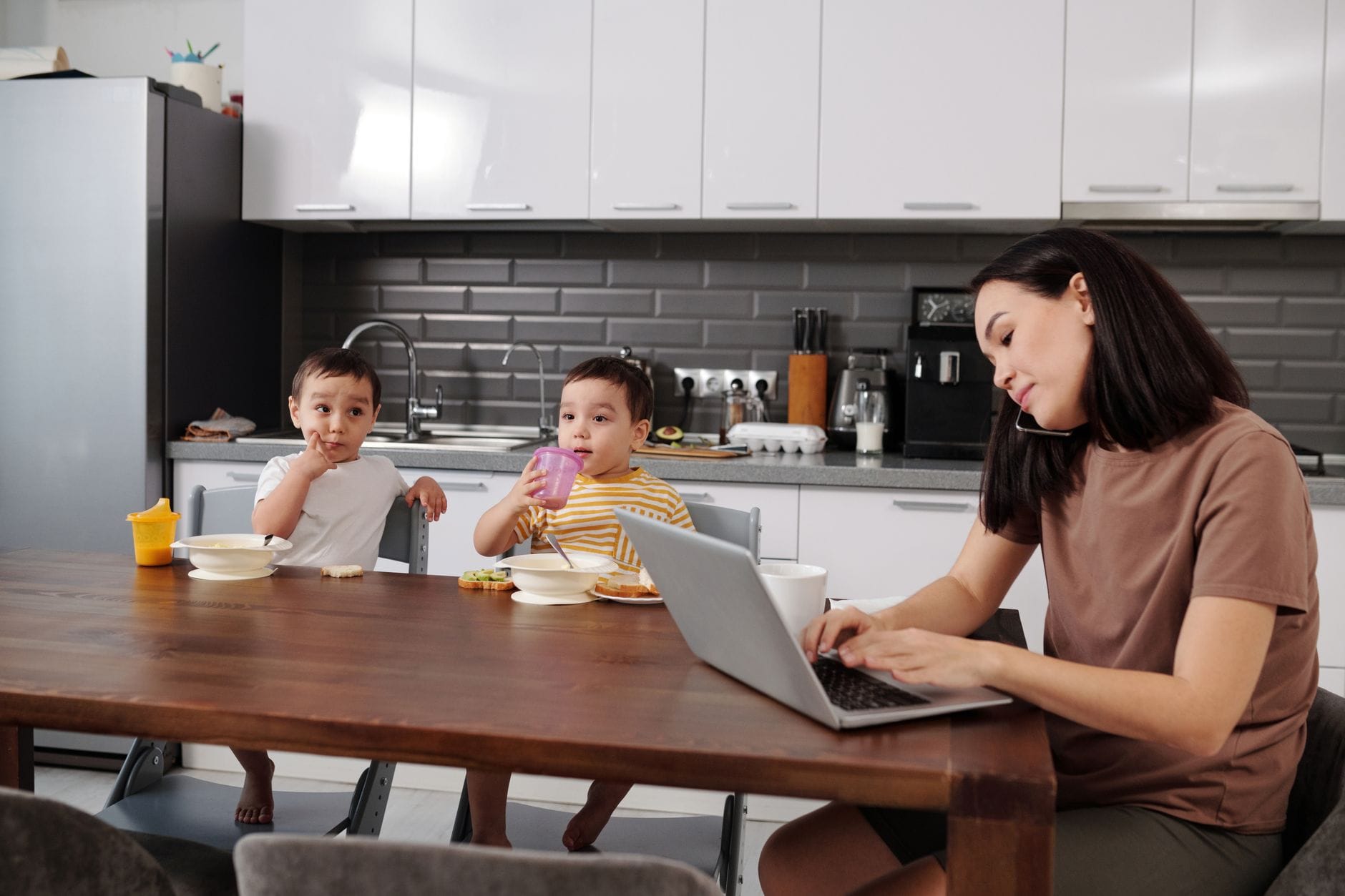 Mama pracuje przy laptopie, a dwaj synowie jedzą śniadanie w kuchni